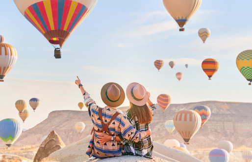 A couple, with arms outstretched, stands united in a picturesque valley in Kapadokya, Anatolia. Above them, hot air balloons take flight, enhancing the beauty of their destination in Goreme, Nevsehir.