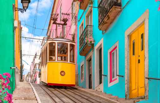 A bright yellow traditional tram traverses a charming street lined with colorful houses adorned with balcony flowers. This picturesque scene unfolds along the Bica Elevator, as it descends the hill of Chiado in Lisbon, Portugal.